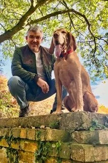 A man with gray hair and glasses crouching next to a large golden retriever dog on a stone wall outdoors under a tree.