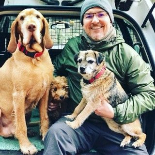 Man in green jacket and gray beanie holding two dogs, one large and one small, in a vehicle with three other dogs.