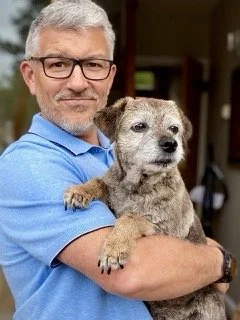 A middle-aged man with glasses and gray hair holding a small, scruffy dog with light brown fur.