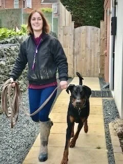 A woman walking a black Doberman puppy on a leash outdoors near a wooden fence and gate.
