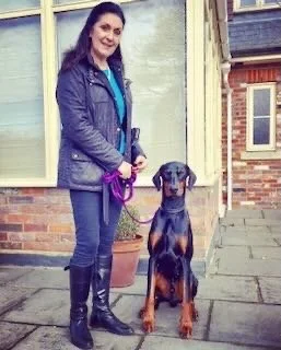 A woman standing outside on a sidewalk with a large Doberman dog sitting beside her, in front of a brick house with large windows.