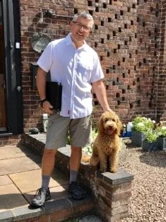 A man stands outside a brick house with a dog on a small brick step, holding a black folder.