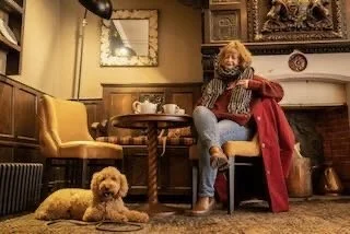A woman with curly hair and a dog sitting on the floor in a cozy, vintage-style living room with a fireplace, mirror, and yellow armchair.