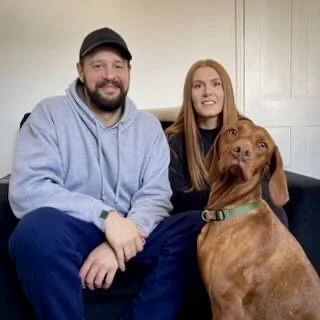 A man with a beard wearing a baseball cap and a woman with long hair sitting on a couch with their brown dog.