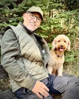 Man sitting outdoors with a curly-haired dog in a wooded area.