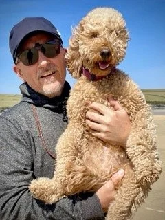 A man wearing sunglasses and a cap holding a fluffy beige dog outdoors against a blue sky.