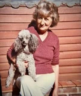 An elderly woman with short curly hair wearing a red sweater and light-colored pants, standing outdoors with a small poodle sitting on her lap, in front of a wooden building.