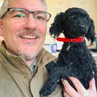 A man with glasses smiling, holding a small black poodle in a red collar.
