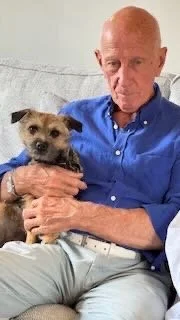 An elderly man wearing a blue shirt sitting on a couch, holding a small dog with tan and black fur.