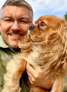 A man smiling while holding a small brown dog outside on a bright day.