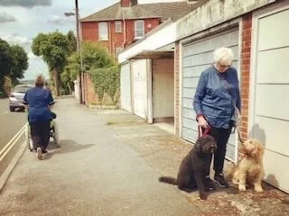 Older woman standing outside with two dogs, one black and one golden-colored, near garages on a residential street.