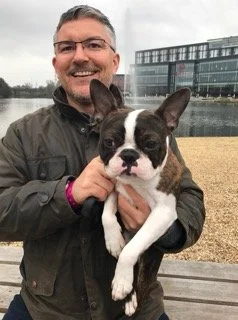 Man holding Boston Terrier dog outdoors near water and modern buildings.