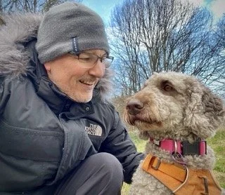 A man wearing glasses, a gray beanie, and a black North Face jacket smiling and looking at a large, curly-haired dog with a pink collar and orange harness outdoors during daytime.