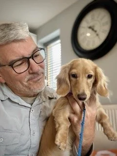 An elderly man with glasses holding a small tan puppy with floppy ears in a room with a wall clock.