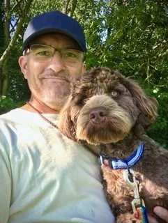 Man wearing glasses and a cap smiling outdoors with a brown and black curly-haired dog in front of him.