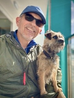 A smiling man wearing sunglasses, a blue cap, and a green jacket, sitting next to a small brown dog with a collar.