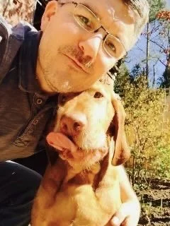 A man with glasses taking a selfie outdoors with a golden retriever puppy, trees and blue sky in the background.