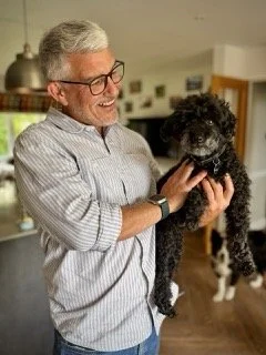 An older man with gray hair and glasses holding a small black poodle inside a home, smiling.