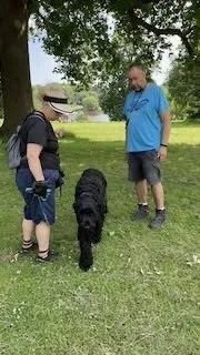 A boy with a backpack, an adult man, and a black dog standing on grass in a park with trees.