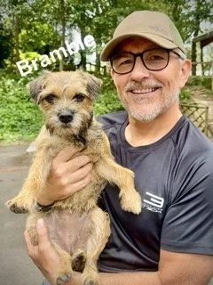 A man in glasses holding a small tan and black dog outdoors with greenery in the background. The word 'Bramble' is written in white text above the dog's head.