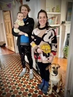 Two women standing in a house with a small dog. The woman on the left is holding a baby, and the woman on the right is wearing a colorful sweater.