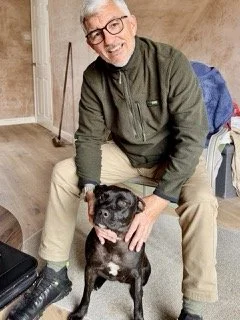 An elderly man with glasses smiling and petting a small black dog sitting on the floor indoors.