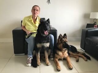 A woman sitting on a black sofa, holding a leash attached to a large black dog sitting on her left and a smaller German Shepherd lying on the floor to her right in a living room.