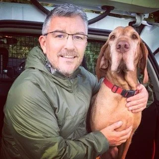 A man with glasses and gray hair smiling while holding a large brown dog with floppy ears and a red collar.