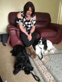 A woman sitting on a maroon leather couch with three dogs—one black and white, one black, and one tan and black—on a patterned rug in front of her.