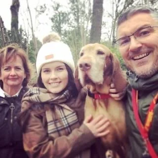 Family of four with a Labrador Retriever outdoors in a forested area, all smiling at the camera.