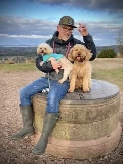 Person sitting on a large circular stone with two dogs, one in a blue sweater and one without, outdoors with cloudy sky and landscape in the background.