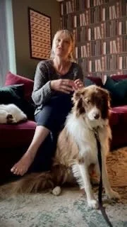 Woman sitting on a sofa with a large dog beside her in a cozy living room filled with books and a chessboard.