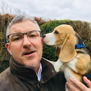 A man with glasses and a beard holding a small beagle puppy outdoors, with bushes and cloudy sky in the background.
