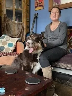 A woman sitting on a couch with a brown and white dog, both looking at the camera. The room has blue walls, a window with curtains, and various decorative items.