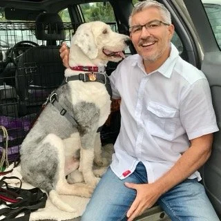 A smiling man in a white shirt sitting in a car with a large, adorable white and gray dog, possibly a husky mix, sitting beside him.