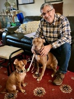 A smiling man sitting on a couch with two dogs, one large and one small, in a living room.