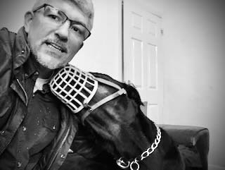 A man with glasses taking a selfie with a large dog wearing a patterned muzzle and a chain collar, indoors.