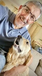 A smiling man wearing glasses and a blue shirt taking a selfie with a golden retriever dog indoors.