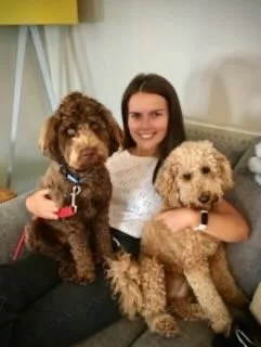 A woman sitting on a couch, smiling, holding two curly-haired dogs, one in each arm.