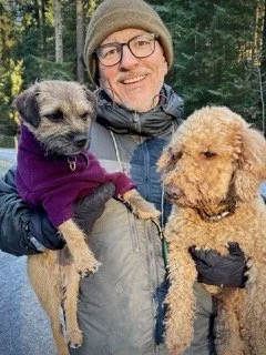 A man in outdoor clothing and a beanie holds two dogs, one on each hand, in a forest setting.