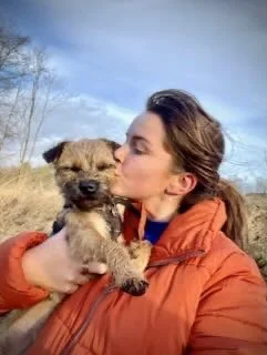 A woman in an orange jacket is holding and kissing a small, fluffy puppy outdoors with a clear sky and trees in the background.