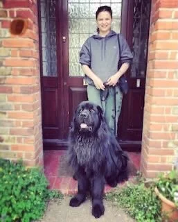 A woman standing in front of a brick house with a large black dog on a leash.