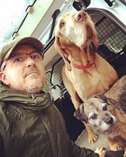 A man wearing glasses and a hat taking a selfie with two dogs inside a dog crate. One dog is large, brown with a red collar, and the other is small, gray and white.
