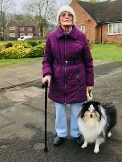 An elderly woman in a purple coat and white hat standing outside on a sidewalk, holding a black cane and a small fluffy black and white dog.