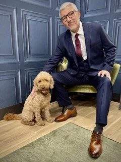 An older man in a dark suit, white shirt, and maroon tie sitting on a green chair, with a fluffy cream-colored dog sitting next to him, in a room with blue paneled walls.