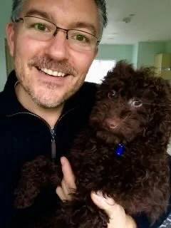 A smiling man wearing glasses holding a small, dark brown dog indoors.