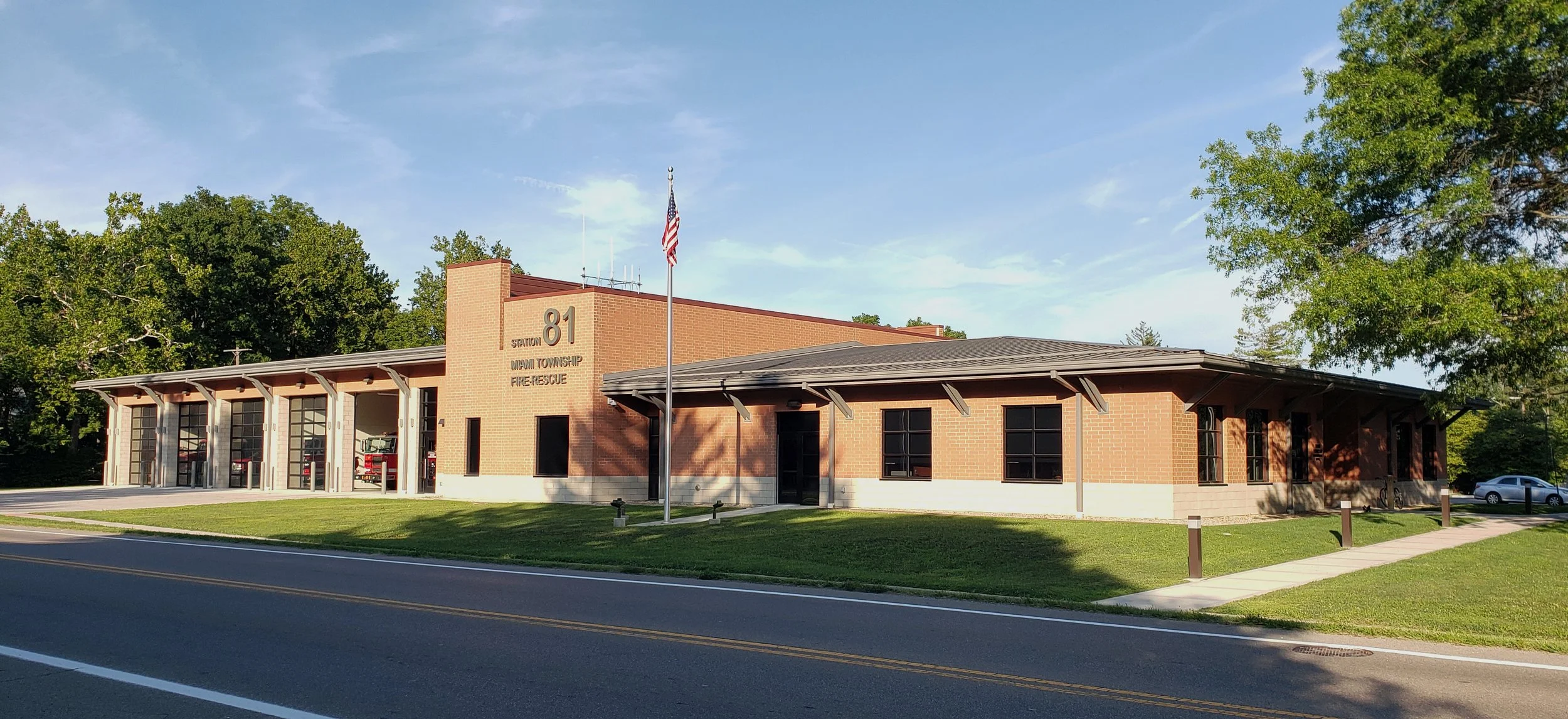 Fire station building labeled 'Station 81 Miami Township Fire-Rescue' with a flagpole and American flag in front, set against a blue sky with trees and parked cars nearby.