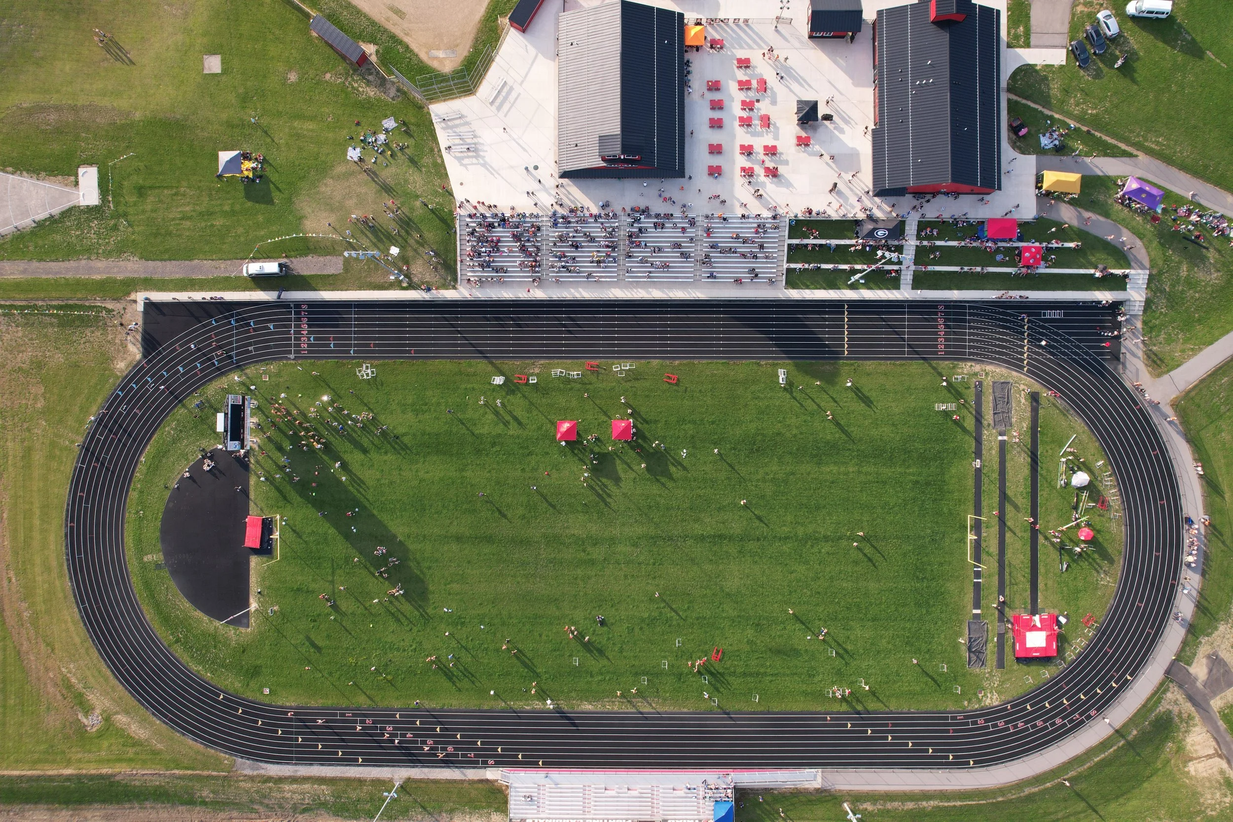 Triad Local School's Track and Field. Aerial view of a sports stadium with a running track, a grassy field, and spectators in the stands. There are tents, vendors, and parking areas surrounding the stadium.