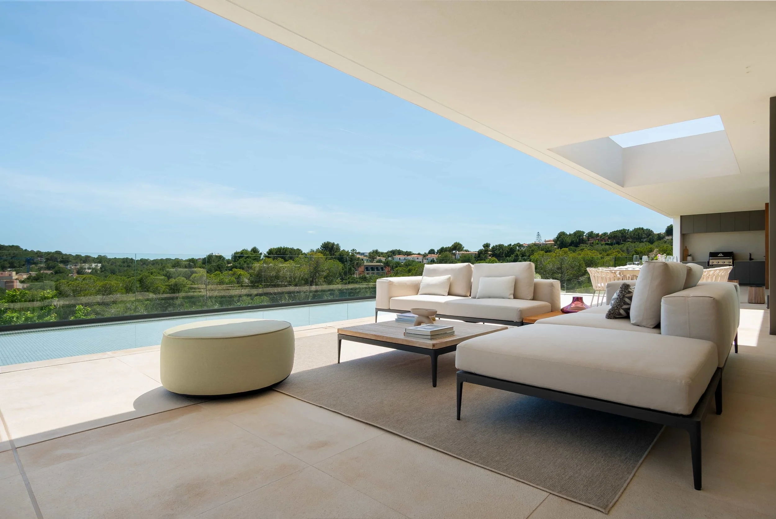 Modern outdoor patio with white cushioned sofas, a round ottoman, a coffee table with magazines, and a dining area with a barbecue grill, overlooking a landscape of green trees and residential houses under a partly cloudy sky.