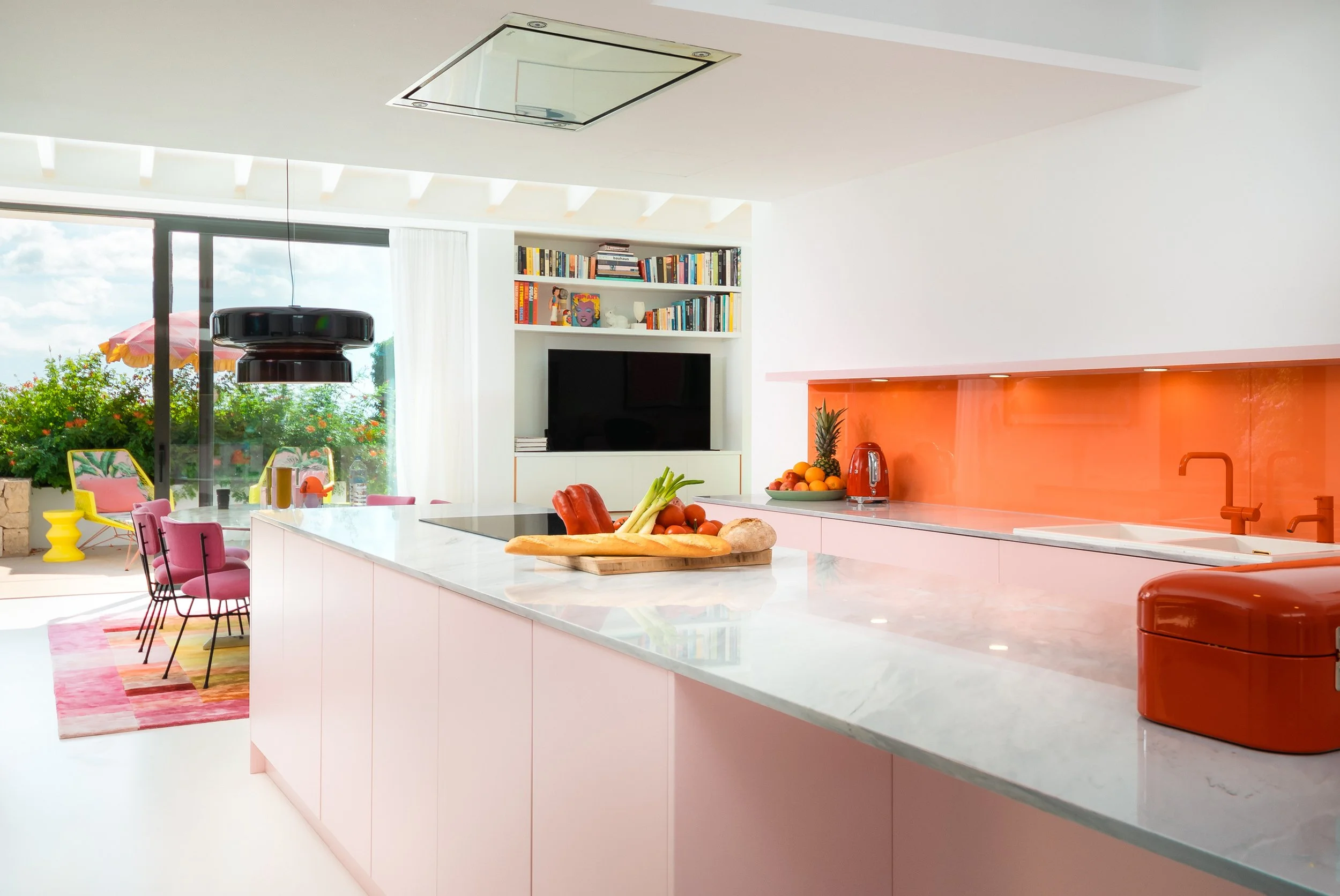 Modern kitchen with pink cabinetry, white marble countertops, orange backsplash, and kitchen appliances. There is a fruit tray with pineapple, oranges, and other fruits, as well as a cutting board with vegetables. In the background, a living area has bookshelves, a television, and large patio doors leading outside where chairs and an umbrella are visible.